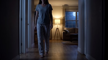 Una mujer en pijama y descalza camina por un pasillo interior oscuro. Al fondo se ve un salón iluminado por una lámpara y una ventana con luna llena.
