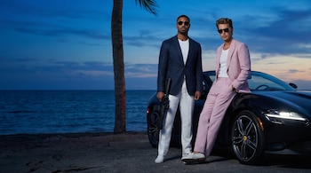 Michael B. Jordan, vestindo terno azul e arma, e Austin Butler, vestindo terno rosa, usando óculos escuros na frente de um carro esporte preto e o oceano ao entardecer.