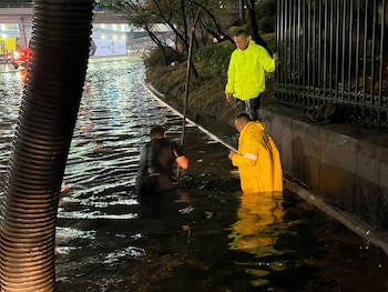 Tres hombres en calle inundada de noche. Uno semisumergido, otro en impermeable amarillo en el agua, y el tercero en chaqueta neón junto a valla. Manguera negra en primer plano