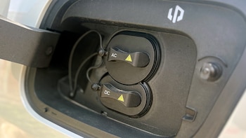 Tapa abierta del puerto de carga de un automóvil con dos cubiertas protectoras marcadas como AC y DC con símbolos de advertencia