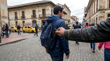 Mano sacando un celular de una mochila azul en una calle adoquinada de Bogotá, con peatones, taxis amarillos y edificios históricos al fondo.