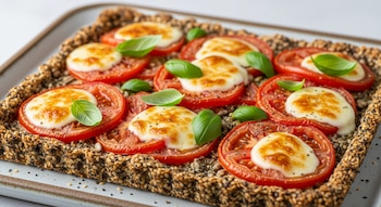 Tarta rectangular con base de semillas, rodajas de tomate, mozzarella fundida y hojas de albahaca, servida en una bandeja de cerámica.