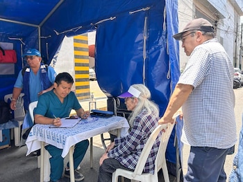 Un profesional de salud escribe sobre una mesa bajo una carpa azul mientras atiende a una mujer sentada frente a él
