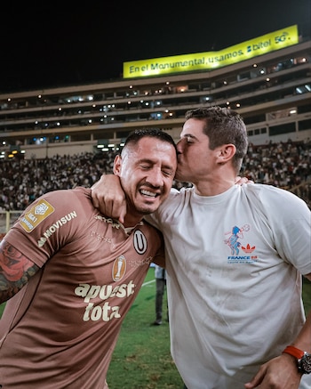 Dos hombres se abrazan en un campo de fútbol. Uno lleva camiseta marrón de equipo, el otro una blanca. Un estadio con público y un cartel luminoso al fondo