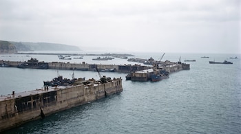 Grandes estructuras de hormigón alineadas en el mar forman un dique junto a barcos y grúas bajo un cielo nublado.