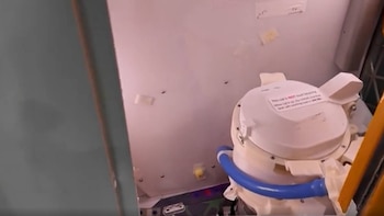 Un inodoro espacial blanco con una tubería azul visible en el interior de una nave espacial. La tapa tiene una etiqueta de advertencia