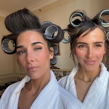 Dos mujeres, Juana Viale y Ámbar de Benedictis, llevan rulos metálicos en el cabello y batas blancas, sonriendo a la cámara en un primer plano