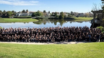 Numeroso grupo de personas con camisetas oscuras posando en un jardín con césped verde, lago, árboles, puente y casas al fondo