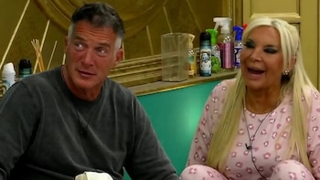 Hombre de pelo gris en suéter oscuro y mujer rubia con pijama rosa estampado, riendo. Sentados en una habitación, con estantes y productos de fondo