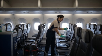 Una mujer uniformada con guantes verdes rocía un desinfectante sobre la bandeja de un asiento de avión vacío. Filas de asientos, ventanillas y equipo de limpieza son visibles.