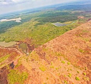 Vista aérea de un paisaje con una línea diagonal que divide campos verdes con cuerpos de agua a la izquierda y tierra rojiza más seca a la derecha bajo un cielo nublado