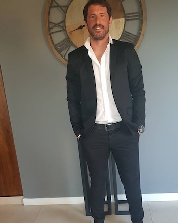Un hombre con traje oscuro y camisa blanca sonríe con las manos en los bolsillos, frente a una pared gris con un reloj de madera grande