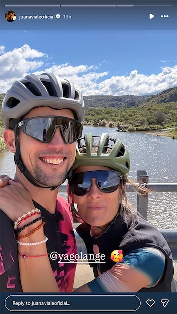Una pareja sonriente con cascos y gafas de sol, Juana Viale y Yago Lange, posa muy cerca frente a un río y colinas verdes bajo un cielo azul con nubes