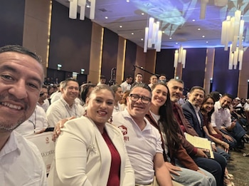 Un hombre con barba tomando una selfi con cinco personas sonrientes, con más asistentes de fondo sentados en un salón de convención con luces colgantes