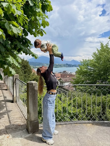 Jimena Barón levanta a su hijo Matías Palleiro en el aire sobre un balcón con barandales de metal, con un lago y montañas de Suiza al fondo