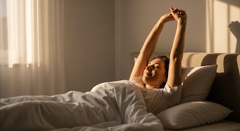Una mujer en la cama se estira con los brazos levantados. Luz natural entra por una ventana en el costado izquierdo e ilumina la habitación.