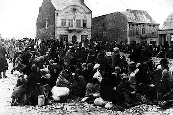 Imagen en blanco y negro de cientos de personas sentadas o de pie en una plaza de tierra. Al fondo se ven edificios europeos de dos y tres pisos con ventanas. Hay bultos y enseres dispersos