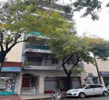 Vista frontal de un edificio de apartamentos de varios pisos, con árboles verdes. Un coche blanco hatchback y una motocicleta están estacionados en la calle