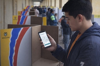 Un joven de perfil con una chaqueta azul y camiseta naranja, mira su celular mostrando un mapa de una app de transporte. Al fondo, se ven puestos de votación.