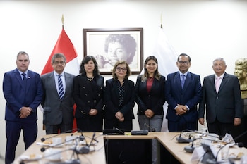 Siete personas con vestimenta formal posan de pie detrás de una mesa de reuniones con micrófonos en un salón con banderas de fondo