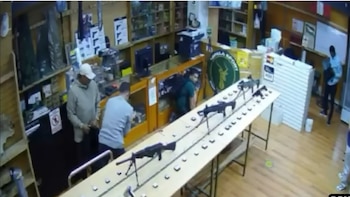Vista aérea de una tienda de armas desde una cámara de seguridad, mostrando varios hombres y fusiles exhibidos sobre un largo mostrador central
