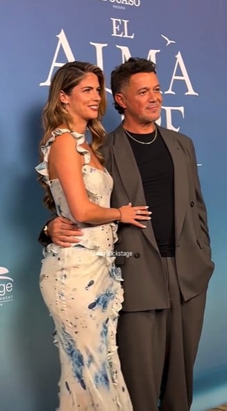 El cantante Alejandro Sanz y la actriz Stephanie Cayo posan abrazados durante un evento público ante un panel azul.