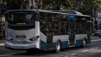 Un autobús plateado, azul y negro de la marca Mercedes-Benz visto desde atrás en una calle con árboles y edificios