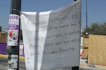Un cartel blanco con texto manuscrito en negro atado a un poste negro. Al fondo, un letrero morado, barreras de madera de construcción y cielo azul