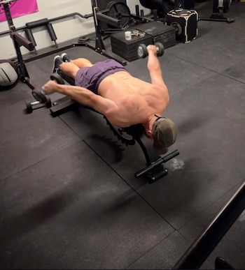 Un hombre sin camiseta boca abajo sobre un banco inclinado levanta dos mancuernas en un gimnasio