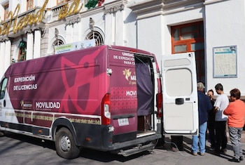 Una van compacta color vino y blanca, con puertas traseras abiertas y el texto 'Expedición de Licencias', está estacionada frente a un edificio con personas alrededor