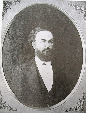 Fotografía en blanco y negro de un hombre con barba y traje oscuro dentro de un marco ovalado con esquinas decoradas
