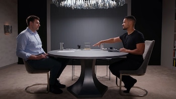 Ben Felix (camisa azul) y un hombre (camiseta negra) sentados en una mesa redonda. La mesa tiene vasos y un objeto transparente etiquetado "$500,000 HOUSE". Fondo moderno