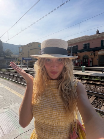 Mujer con cabello rubio, sombrero de paja, vestido amarillo a cuadros en andén de estación de tren con vías, edificios y señal Monterosso