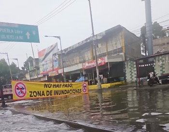 Calle inundada con un gran letrero amarillo que dice "NO HAY PASO, ZONA DE INUNDACIONES". Al fondo se ven edificios y un letrero de carretera