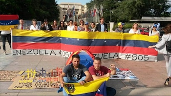 Un grupo de personas sostiene una gran bandera de Venezuela con el texto "VENEZUELA #SOS". Dos hombres se sientan en el suelo con otra bandera más pequeña, y hay pequeñas cruces y pancartas