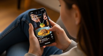 Vista superior de una persona sentada, sosteniendo un smartphone negro en vertical con ambas manos, viendo un reel de comida que muestra un plato de pasta.