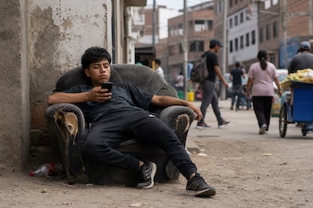 Un joven sentado en un sillón deteriorado en la calle mira su teléfono celular mientras personas caminan al fondo.