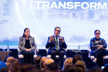 Dos mujeres y un hombre sentados en sillas blancas en un escenario. El hombre sujeta un micrófono. Pantalla de fondo con texto "TRANSFORMA". Botellas de agua. Audiencia borrosa