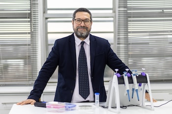 Un hombre con anteojos, saco y corbata posa apoyado en una mesa de laboratorio con pipetas y tubos delante de una ventana con persianas