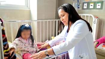 Una médica vestida con bata blanca se inclina hacia una niña sentada en una cama de hospital con barandas