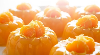 Varias gelatinas translúcidas de color naranja suave en moldes individuales, decoradas con gajos de mandarina y ralladura de jengibre, sobre un fondo blanco luminoso.