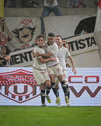 Tres futbolistas de Universitario con uniformes blancos celebran un gol en un campo verde por la noche, abrazándose ante una pantalla LED y pancartas del estadio