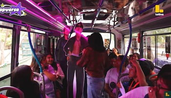 Interior de un autobús con luces de neón moradas, pasajeros sentados, siluetas de cartón y una persona con micrófono en el pasillo
