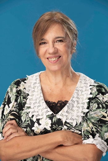 Ingris Pelicori sonriendo, cabello castaño claro, top floral negro con cuello de encaje blanco. Tiene los brazos cruzados y un fondo azul detrás