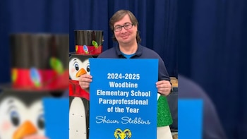 Hombre caucásico con gafas y polo azul sonríe a la cámara, sosteniendo un cartel azul que lo nombra Paraprofesional del Año de la Escuela Primaria Woodbine