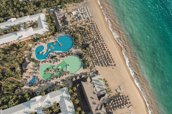 Vista aérea de un complejo hotelero con piscinas de formas irregulares, palmeras, sombrillas en la arena y la orilla del mar turquesa