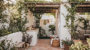 Un patio con pérgola de madera y enredaderas, paredes blancas y suelo de baldosas. Hay una lavadora, fregadero, ropa tendida, plantas, cestos y un sofá.
