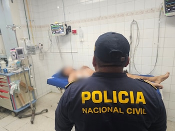Vista trasera de un oficial de Policía Nacional Civil con gorra, frente a un paciente borroso en camilla, rodeado de equipo médico en una sala con paredes de azulejos