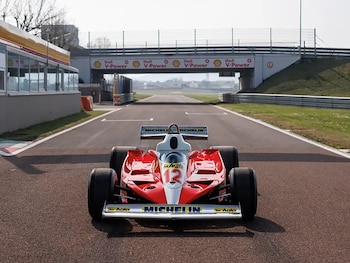 Coche de Fórmula 1 Ferrari 312 T3 rojo y blanco con el número 12 estacionado en una pista de carreras. Un puente con logotipos de Shell V-Power está al fondo