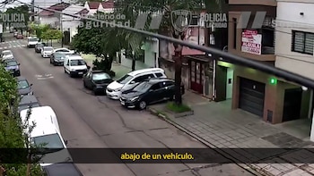 Calle residencial con automóviles estacionados, aceras y casas, registrada por una cámara de vigilancia con logotipos institucionales